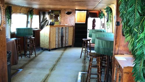 Interior of a vintage bus converted into a lounge, featuring wooden decor, bar area, and tropical plants.