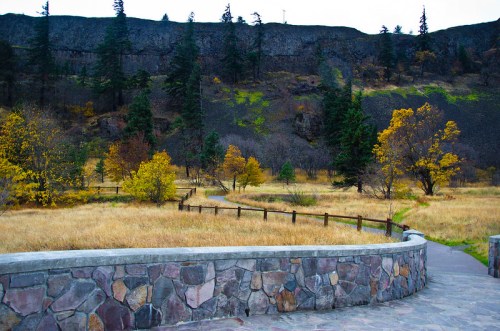 A scenic view of a grassy area with trees and a stone pathway, set against a rocky hillside in autumn.