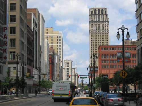 A busy city street with tall buildings, a bus, and a taxi under a partly cloudy sky.