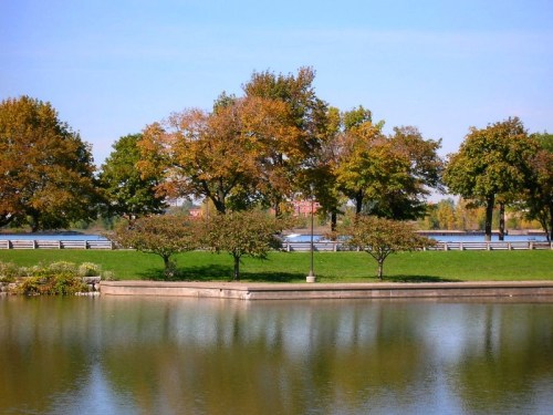 A serene park scene with trees in autumn colors reflected in a calm lake under a clear blue sky.