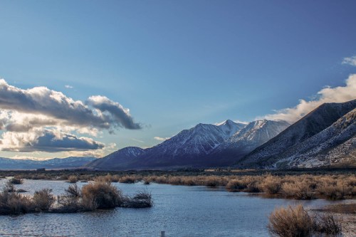Scenic view of mountains with snow, reflecting in a calm lake surrounded by shrubs and a cloudy sky.