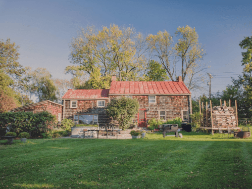 A rustic house with a red roof, surrounded by greenery and a neatly maintained lawn, with firewood stacked nearby.