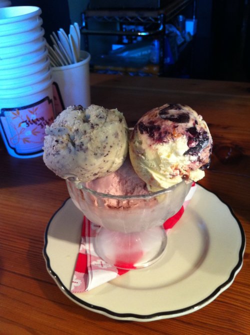 A glass dish holds three scoops of ice cream: cookies and cream, strawberry, and a berry swirl, on a wooden table.