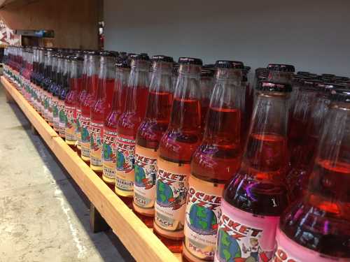 A row of colorful soda bottles on a wooden shelf, featuring vibrant labels and various flavors.