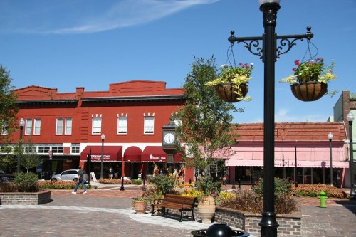 A vibrant town square with red buildings, hanging flower baskets, and people walking on a sunny day.