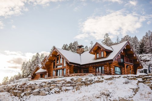 A large log cabin with a snow-covered roof, surrounded by trees and a rocky landscape under a cloudy sky.