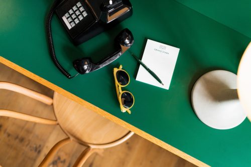 A green desk with a black phone, yellow sunglasses, a notepad, and a pen, beside a lamp and a wooden chair.