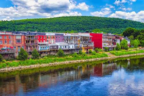 Colorful buildings line a riverbank, reflecting in the water, with a lush green hillside in the background under a blue sky.