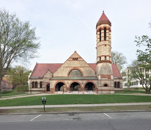 Historic building with a red and stone facade, featuring a tall tower and arched entrances, surrounded by green grass.
