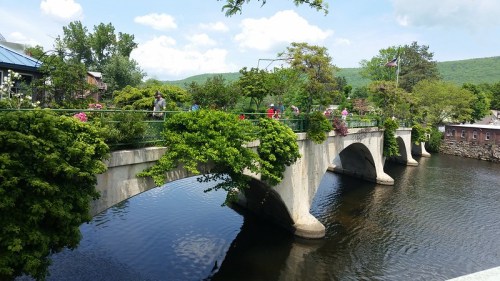 A scenic view of a stone bridge over a calm river, surrounded by greenery and people enjoying the outdoors.