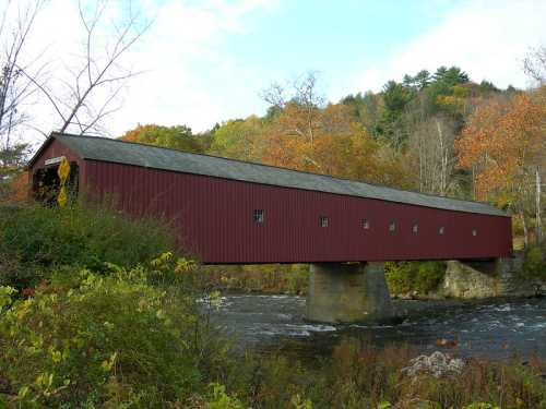 A red covered bridge spans a river, surrounded by autumn foliage and trees under a partly cloudy sky.