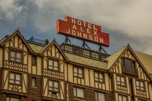 Sign for Hotel Alex Johnson atop a historic building with a mix of architectural styles under a cloudy sky.