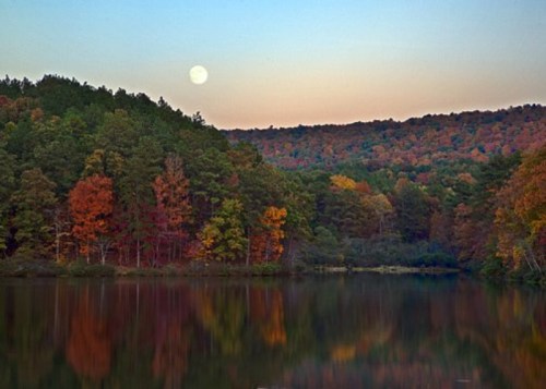 A serene lake reflects colorful autumn trees and a full moon against a twilight sky.