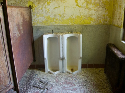 Two old, worn urinals in a dilapidated restroom with peeling yellow walls and debris on the floor.
