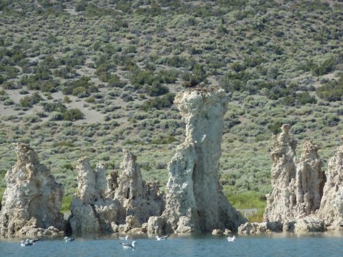 Rock formations rise from a calm lake, surrounded by green hills and a few birds swimming nearby.
