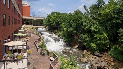 A scenic view of a river with waterfalls, surrounded by greenery and a patio area with tables and umbrellas.