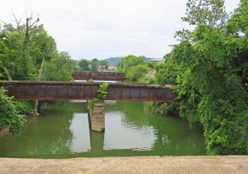 Two old railway bridges cross a green river, surrounded by lush trees and a cloudy sky.