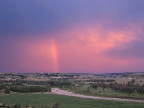 A scenic landscape featuring rolling hills, a river, and a vibrant rainbow against a purple and pink sky.