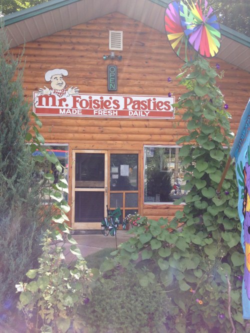 A wooden building with a sign reading "Mr. Poisie's Pasties" and colorful decorations, surrounded by greenery.