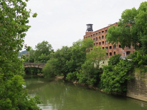 A serene river scene with lush greenery and a historic brick building in the background under a cloudy sky.