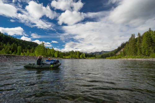 A person in a raft floats on a calm river surrounded by lush green trees and a blue sky with fluffy clouds.