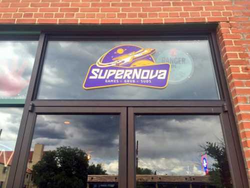 Sign for "Supernova" featuring the name and a graphic of a rocket, displayed on a window with cloudy skies in the background.