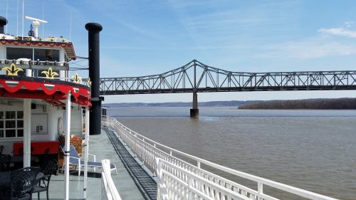 A riverboat docked beside a bridge over a calm river on a sunny day.