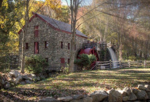 A historic stone mill with a water wheel, surrounded by trees and a grassy area, in a serene outdoor setting.