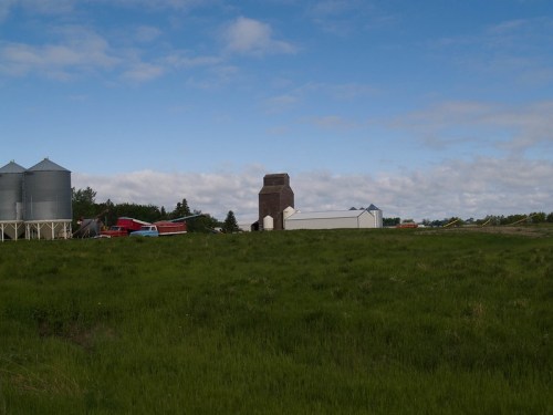 A rural landscape featuring silos, a barn, and a grassy field under a partly cloudy sky.