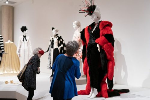 Two women admire elegant fashion displays in a museum, featuring stylish dresses and a striking red fur coat.