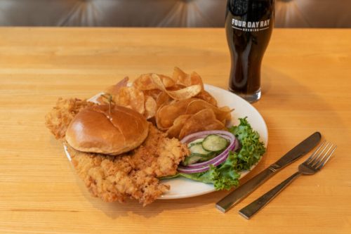A plate with a fried chicken sandwich, chips, lettuce, cucumber, and onion, alongside a dark beer in a glass.