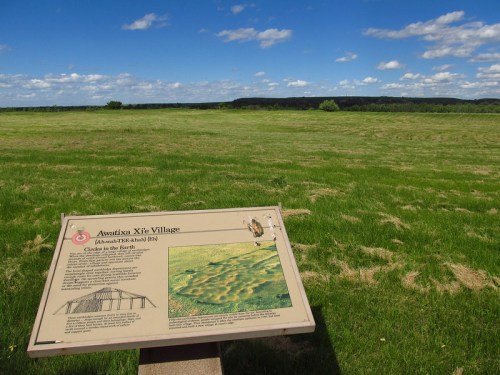 A sign at Awatixa Xite Village with a grassy landscape and blue sky in the background, showcasing historical information.