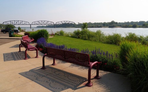 A serene riverside scene with benches, colorful flowers, and a bridge in the background under a clear sky.