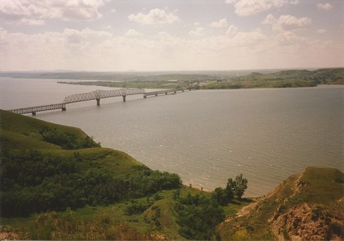 A scenic view of a river with a bridge, surrounded by green hills and a cloudy sky.