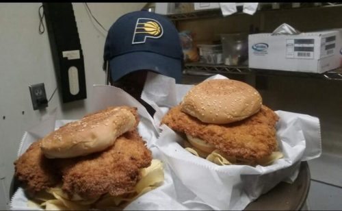 Two large fried chicken sandwiches on buns, served in baskets, with a person wearing a blue cap in the background.