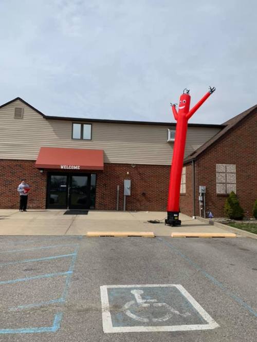 A tall, red inflatable tube man waves outside a building with a "Welcome" sign, while a person stands nearby.