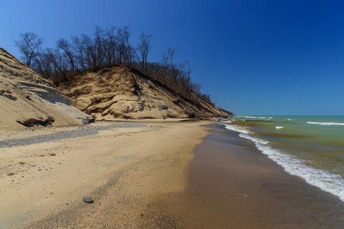 A sandy beach with gentle waves, bordered by a steep, tree-covered hill under a clear blue sky.