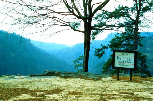 Sign for Petit Jean State Park in Arkansas, with a scenic view of mountains and trees in the background.