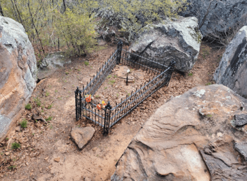 A small, fenced grave surrounded by large rocks and sparse vegetation in a natural setting.
