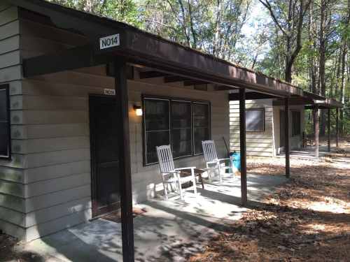 A small cabin with a porch, featuring two rocking chairs, surrounded by trees and fallen leaves.