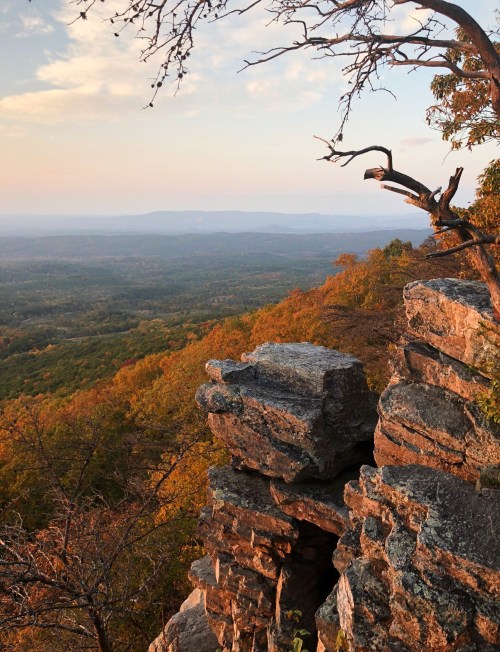 A scenic view from a rocky ledge overlooking a valley filled with autumn foliage and distant mountains.