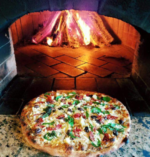 A freshly baked pizza with toppings, placed in front of a wood-fired oven with flames in the background.