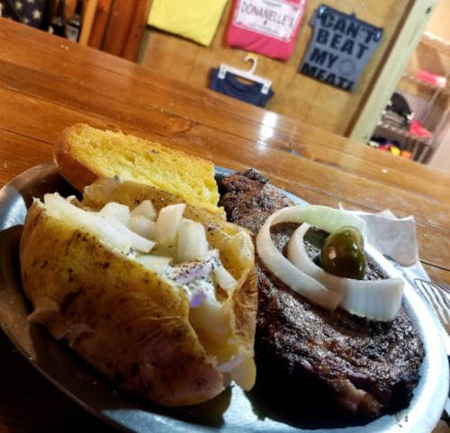 A plate with a baked potato topped with onions, a grilled steak, and a slice of cornbread, set on a wooden table.