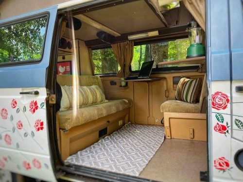 Cozy van interior with floral decor, seating area, and a small desk with a laptop, surrounded by greenery.