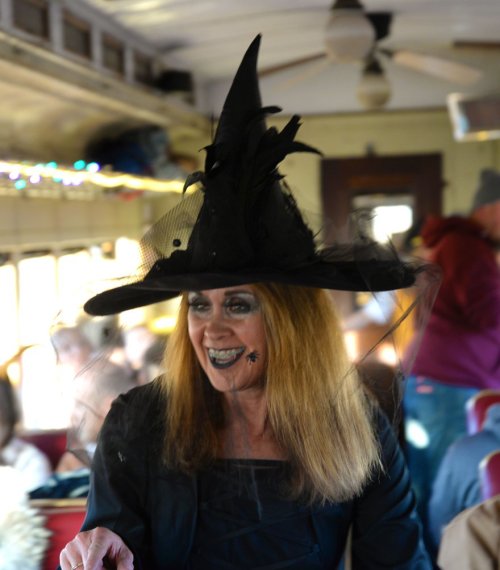 A person in a large black witch hat and dark attire smiles, surrounded by a festive atmosphere on a bus.