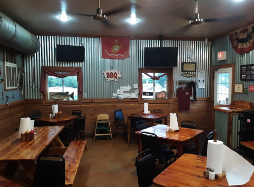 Interior of a rustic BBQ restaurant with wooden tables, metal walls, and decor including flags and photos.