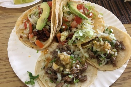 A plate of three tacos filled with meat, lettuce, tomatoes, avocado, and onions, served with lime wedges.