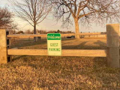 A wooden fence with a "Welcome Guest Parking" sign in a grassy area, with trees in the background.