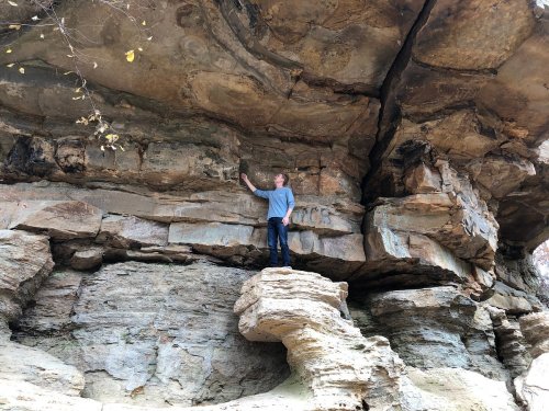 A person stands on a rock ledge, reaching up to touch a rocky overhang in a natural outdoor setting.