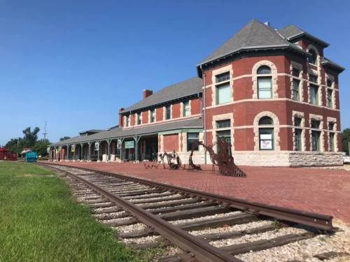 Historic brick train station with a large porch, surrounded by green grass and old railway tracks under a clear blue sky.